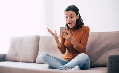 Happy excited beautiful woman with smart phone on the couch