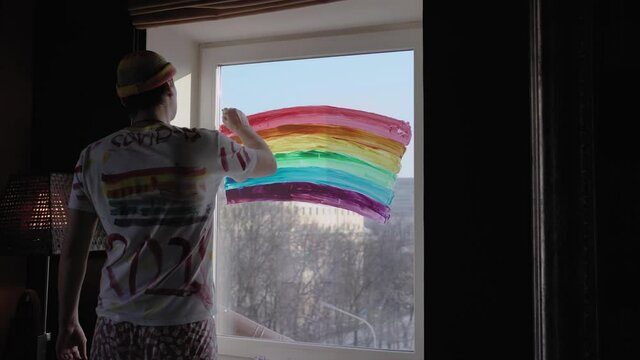 An unusual guy freak draws a rainbow of six colors on his window of the house like an LGBT flag, on it a painted T-shirt, self-isolation at home in the evening.