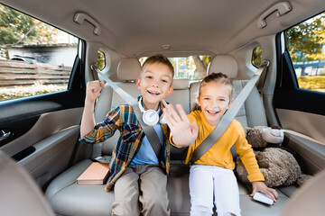 Smiling kids showing yeah and waving at camera near smartphone and book in car
