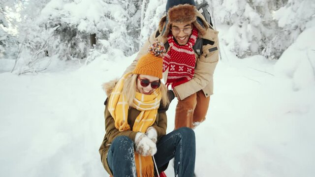 Young man pushing his happy girlfriend on sled through snowy forest on winter day and then falling down and laughing