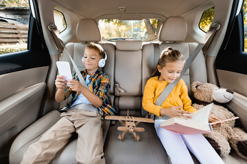 Smiling kids with smartphone and book sitting near toys on back seat of car