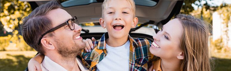 Cheerful son embracing parents near car on blurred background outdoors, banner