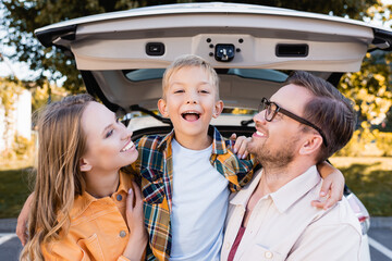 Smiling parents looking at child near auto on blurred background outdoors