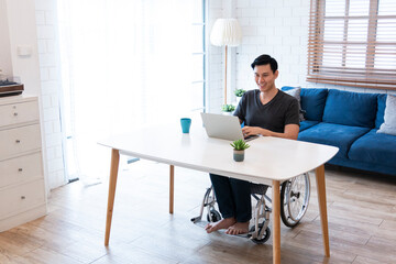 Happy young Man in wheelchair work in his own company from home. Cheerful businessman smile while sitting in his wheelchair. Disabled Man In Wheelchair Using Laptop At Home.