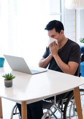 Sick Business entrepreneur in wheelchair sneezing in the home office. Disabled Sick Young Man on Wheelchair at Home. Young man in wheelchair blowing his nose in a tissue at his home office.