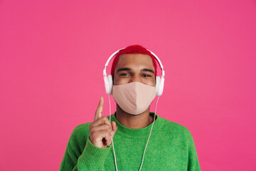 Joyful african guy in face mask and headphones pointing finger upward