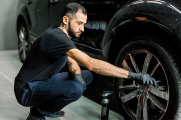 Close up of handsome young male employee worker cleaning the wheel rim of modern black crossover...