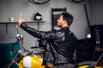 A young biker in a leather jacket in a garage smiles for a photo and his blog