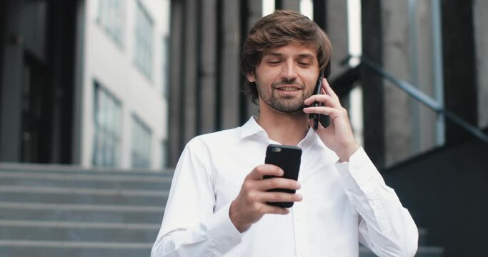Young Successful Businessman with Long Hair is working outdoor, holding two Smartphones. Hipster Successful Businessman is talking, smiling, standing outdoor near Modern Office. Busy Lifestyle.