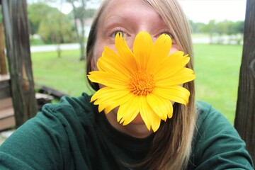 A young blond woman with her face covered by a big yellow flower, tithonia.