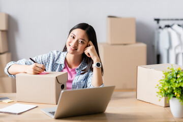 Smiling asian manager of charity center writing on box with sticky note near laptop on blurred foreground