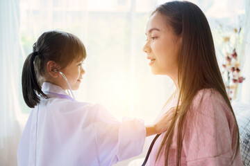 Cute little girl as doctor playing with happy mom pretending nurse, playful kid daughter dressed as medical worker have fun holding stethoscope listening to mother chest, funny child mum game at home