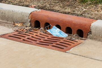 Closeup of surgical face mask discarded in storm sewer drain grate. Concept of Covid-19 coronavirus pandemic pollution and safe face covering disposal.