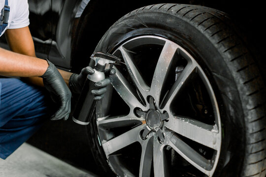 Washing a car by hand, car detailing. Cropped image of hands of young African man in protective gloves spraying special cleaning solution on the rim of modern car at carwash service