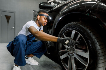 Washing a car by hand, car detailing. Close up image of professional African male worker, cleaning the car wheels with brush. Car rims wash, manual wheel cleaning concept.