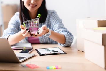 Cropped view of woman showing shopping cart while sitting at desk with devices and carton boxes on blurred foreground
