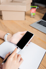 Cropped view of woman holding smartphone near notebook at desk with carton boxes on blurred background