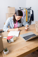 Asian volunteer with smartphone writing in notebook at desk with laptop, carton boxes and scotch dispenser on blurred background