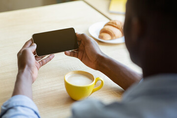 Unrecognizable African American guy holding mobile phone with empty screen at table in cafe, mockup for design