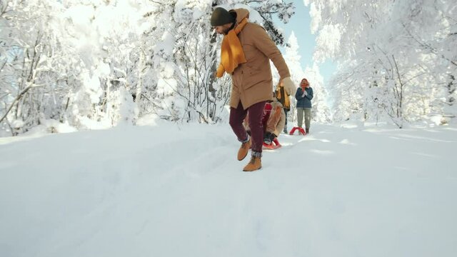 Friends having fun in winter forest: young man running down the hill and pulling sled with excited male friend riding it while their girlfriends jumping and clapping hands