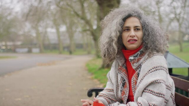 Youthful looking woman with grey hair talking to someone off camera as she sits on a  park bench 