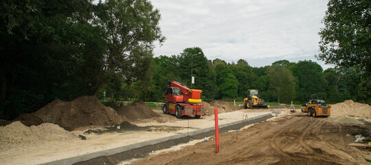 Construction site of new road and sidewalk in city. Yellow tractor and road roller. Green trees in suburban.