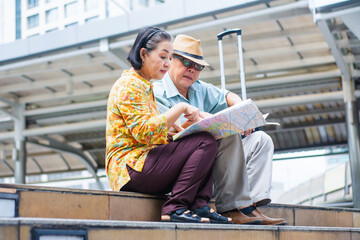 Elderly Asian couples,tourists sitting looking at map showing routes in capital of business districts and planning travel plan with concept of traveling around the world after retirement from work.