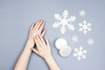 Female hands applying moisturizing cream on skin. Jar with cream, snowflakes flat lay on blue background top view. Winter skincare, skin protection cosmetic. Body, face spa product.