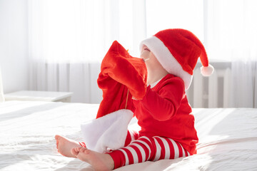 toddler boy in Santa hat, red suit with Christmas sock sitting on white bed at home. sunny morning.