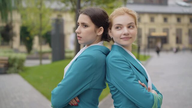 Elegant young Caucasian women in stewardess uniform standing back to back with crossed hands and smiling. Side view portrait of confident beautiful air hostesses outdoors on city street.