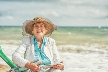 Fototapeta premium Happy laughing old woman 84 years old sitting on the seashore with a tablet computer in her hands.