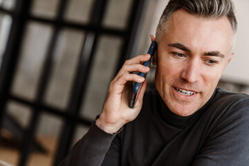 Handsome pleased man talking on mobile phone while sitting at home