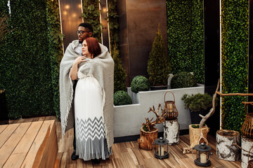 Beautiful couple wrapped in blanket stand at the balcony, handsome African American man hug gorgeous white woman, enjoy tender moments, winter holidays, Christmas morning concept