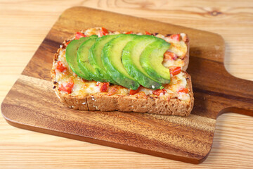 Closeup a homemade grilled cheese toast topped with tomato and sliced avocado on wooden Breadboard