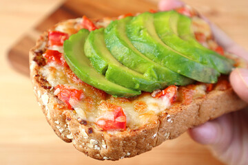 Closeup a mouthwatering grilled cheese toast with tomato and sliced fresh avocado in hand	