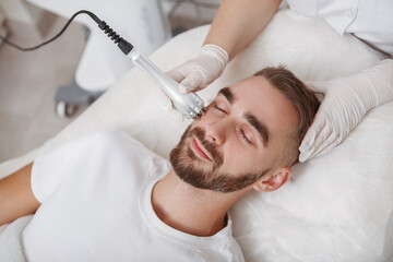 Obraz na płótnie Canvas Attractive bearded young man enjoying hardware cosmetology facial treatment