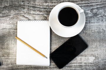 on the wooden table is a Notepad with a pencil a white Cup of coffee and a mobile phone.