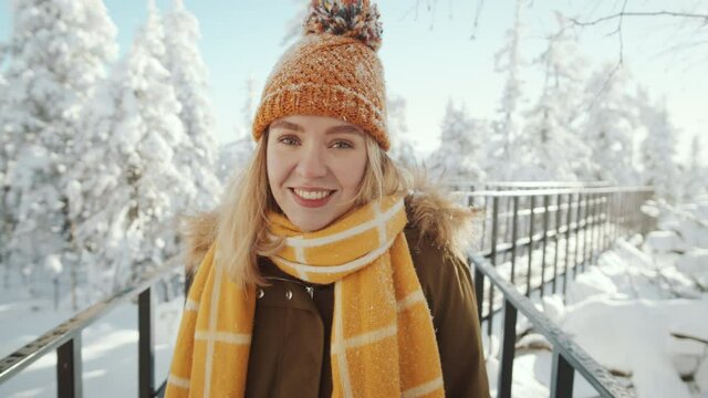 Zoom in portrait shot of young pretty woman in outerwear, woolen hat and scarf smiling and posing for camera while standing on boardwalk in national park on winter day