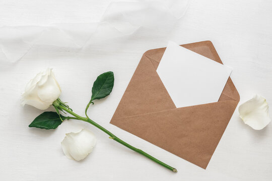 Wedding invitation. Blank card in brown envelope mock-up, white rose and silk ribbon. White wooden painted background. Top view, flat lay.