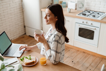 Beautiful smiling woman working with laptop while having breakfast
