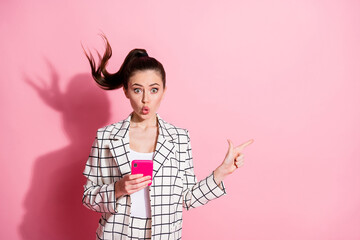 Photo portrait of shocked woman holding phone in one hand pointing finger at blank space isolated on pastel pink colored background