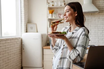 Zelfklevend Fotobehang Zwangere gelukkige vrouw die lacht tijdens het eten van sandwich © Drobot Dean