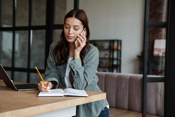 Beautiful serious woman talking on cellphone while working with laptop