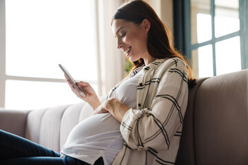 Happy charming pregnant woman using mobile phone while sitting on sofa