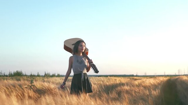 Young hipster girl holding guitar in hand and walking in the wheat field. Woman with acoustic instrument enjoying nature and sunlight. People, lifestyle, student, expression and style concept.