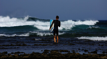 Young man entering the water through the stones to surf, with a surfboard.