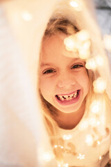 A portrait of a laughing 6-year-old girl hiding in a white plaid and festive garlands.