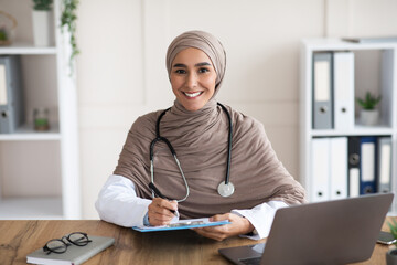Smiling woman doctor in hijab taking anamnesis in clinic - obrazy, fototapety, plakaty