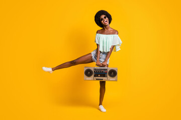 Full size photo portrait of laughing girl standing on one leg dancing holding boombox isolated on vivid yellow colored background