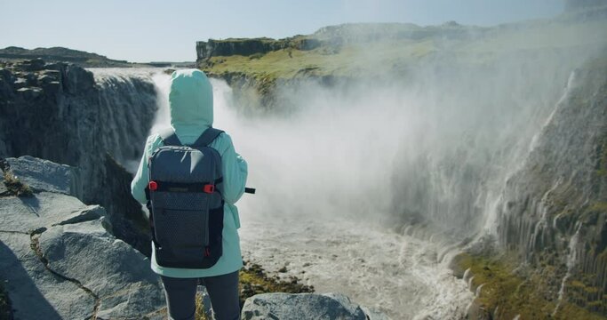 Woman with jacket and backpack standing at cliff edge looking at Detifoss waterfall in Iceland